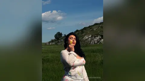 Dreamy woman in a floral white dress holding a yellow flower in a sunny green meadow under a blue sky.