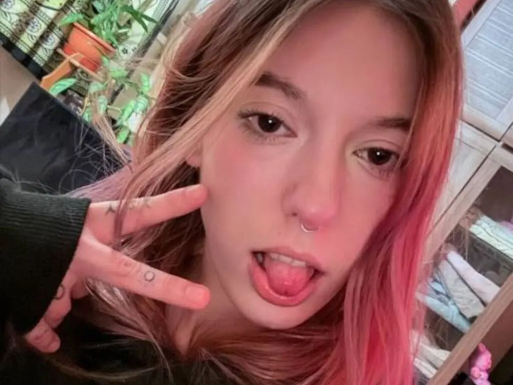 Young woman with pink hair and nose ring making a peace sign in a cozy indoor setting.