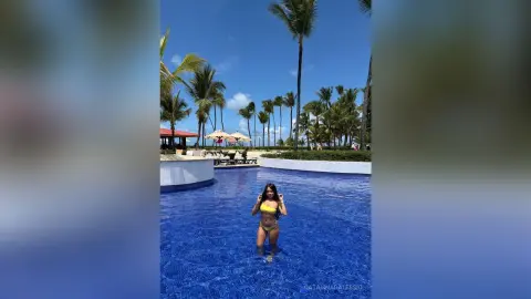 Young woman in a yellow bikini standing in a tropical resort swimming pool under palm trees on a sunny day.