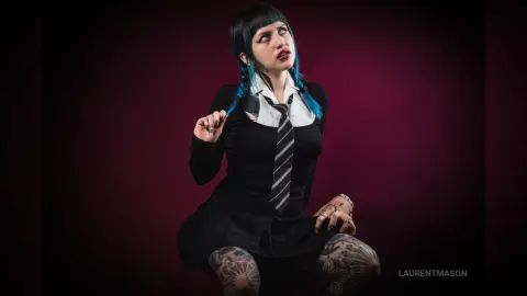 Goth woman with blue hair and tattoos wearing a black dress and striped tie against a dark moody background.