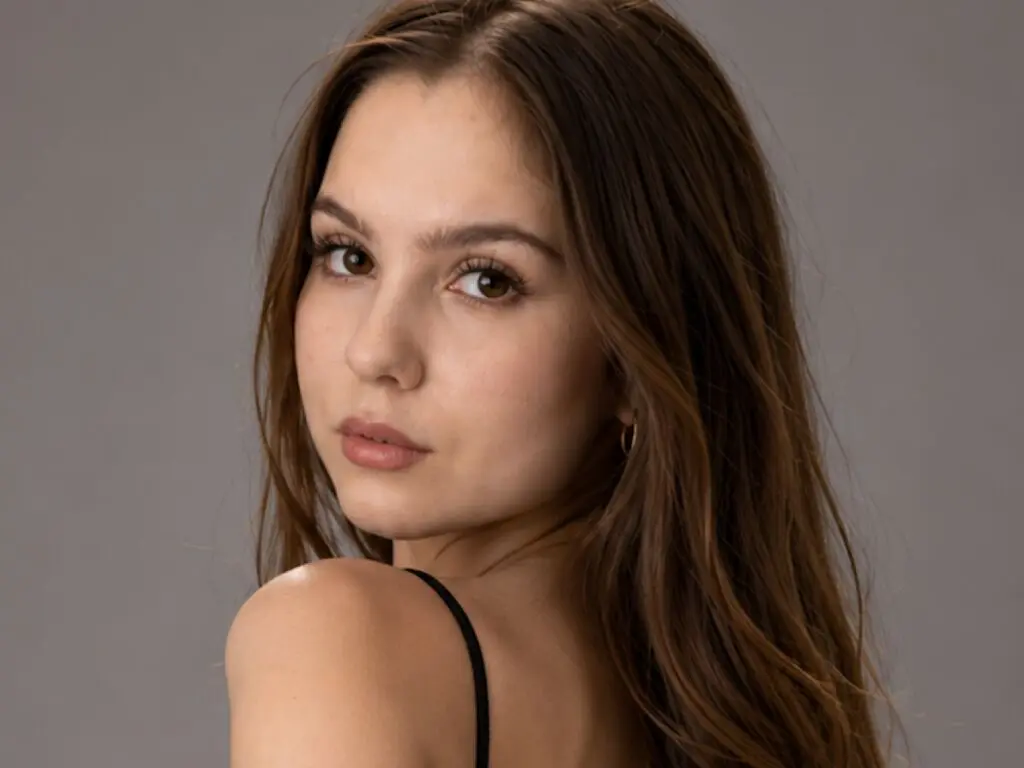 Portrait of a young woman with long brown hair wearing a black strap top against a neutral gray studio background.