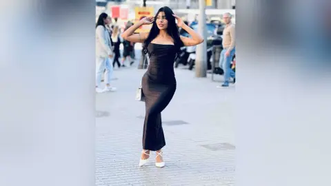 Elegant woman in a sleek black evening gown posing on a sunny city sidewalk.