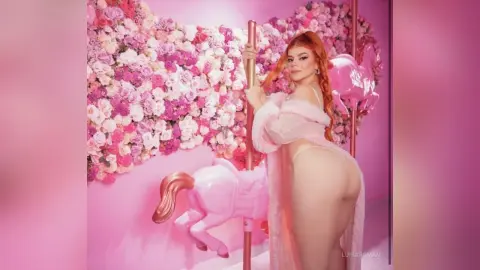 Redheaded woman in a pink fuzzy robe posing against a floral wall and pink unicorn decor in a dreamy setting.