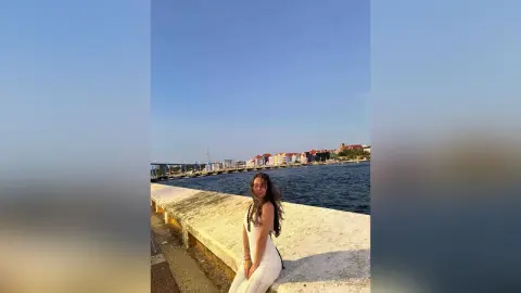 Young woman in a white summer dress sitting on a stone pier by the blue sea against a coastal city skyline.