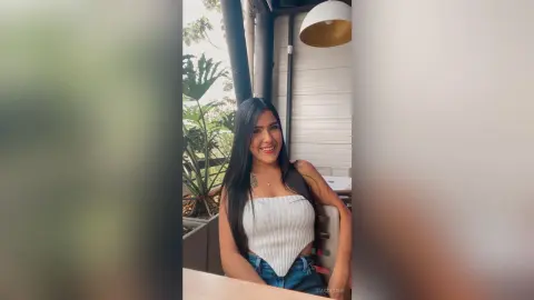 Smiling young woman in a white ribbed tank top sitting at an outdoor cafe patio with lush green plants.