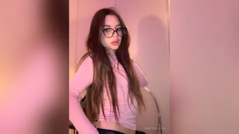 Young woman with long brown hair wearing glasses and a pink long-sleeve top in soft pink lighting.