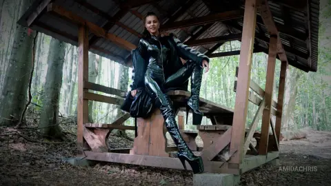 AmidaChris sits on a wooden bench under a shelter wearing a black shiny latex bodysuit and thigh-high platform boots.