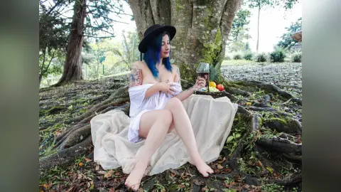 Blue-haired woman in a white wrap sitting on a blanket by tree roots, enjoying wine in a peaceful forest setting.