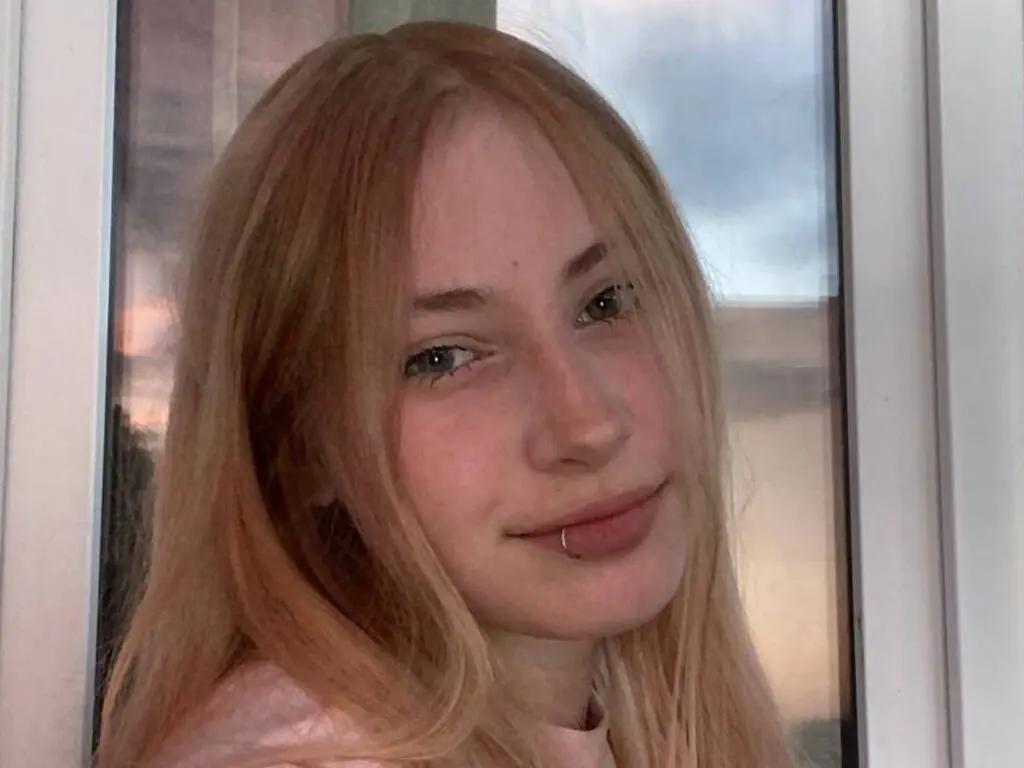 Blonde woman in a pink shirt smiling softly by a window during golden hour.