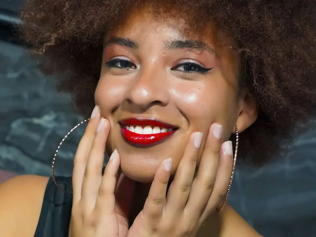 Smiling Black woman with afro hair, red lipstick, and gold hoop earrings in a moody close-up portrait.