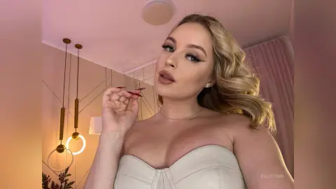 Blonde woman in a white strapless top posing in a warm, soft-lit room with elegant gold pendant lights.
