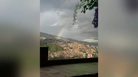 Panoramic view of a sprawling hillside city under a dramatic cloudy sky with a faint rainbow appearing.