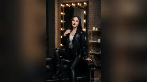 Stylish woman in a black leather jacket and leather pants sitting on a sofa in a moody, dimly lit studio.