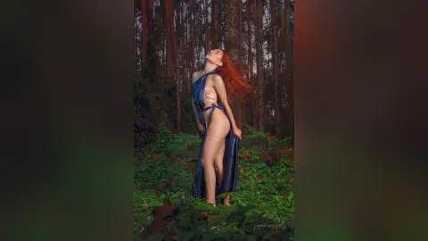 LaurentBelluci stands in a forest wearing a blue open-front robe that exposes her bare breasts, stomach, and pussy.