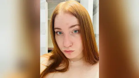Redheaded woman with blue eyes wearing a delicate necklace in a bright bathroom setting.