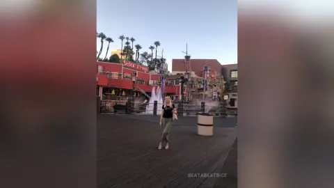 Woman in black top and grey trousers standing on a wooden boardwalk near a fountain at dusk.
