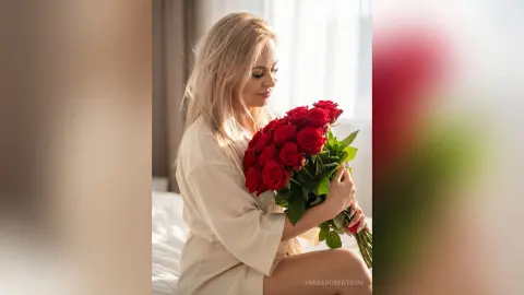 Blonde woman in a cream silk robe holding a large bouquet of red roses in a bright, dreamy bedroom setting.