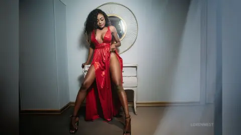 Elegant woman in a glamorous red evening gown posing in a moody, dimly lit room.