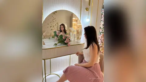 Elegant woman in a white top and pink skirt sitting at a luxury vanity mirror with floral decor.