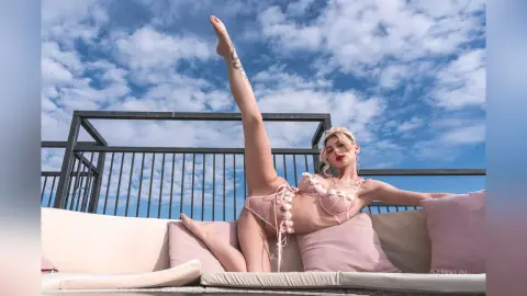 DaisyLin reclines on a light pink outdoor sofa, wearing pink lace lingerie with her right leg raised high against a blue cloudy sky.