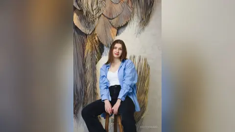 Smiling young woman in a blue striped shirt and black pants sitting against a textured leaf wall decor.