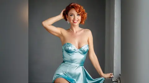 Smiling redhead woman in a light blue satin strapless dress posing in a studio setting.