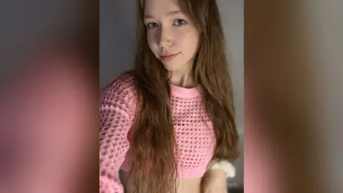 Young woman with long brown hair wearing a pink crochet crop top in a soft, dreamy indoor setting.