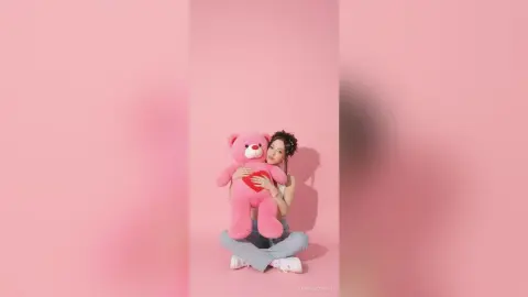 Young woman in casual jeans hugging a large pink teddy bear against a soft pink background.