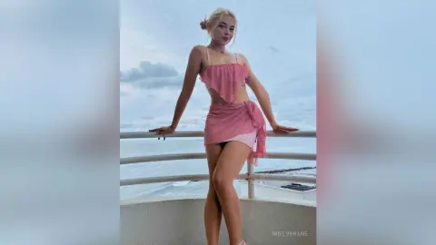 MielVerane poses on a balcony overlooking the ocean, wearing a pink two-piece outfit that shows her midriff and legs.