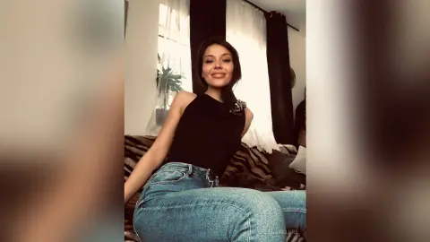 Smiling woman in a black sleeveless top and blue jeans sitting on a sofa in a bright living room.