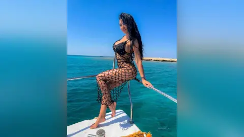 NicoleBella poses naked in a black fishnet bodysuit while sitting on a boat railing overlooking the blue ocean.