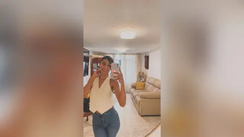 LaraOlson smiling in a white sleeveless top and blue jeans, taking a mirror selfie in a bright living room.