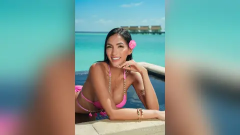 Smiling woman in a pink bikini relaxing by a tropical turquoise ocean resort pool.