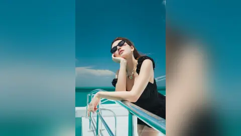 Stylish woman in black summer outfit and sunglasses relaxing on a boat under a clear blue sky.