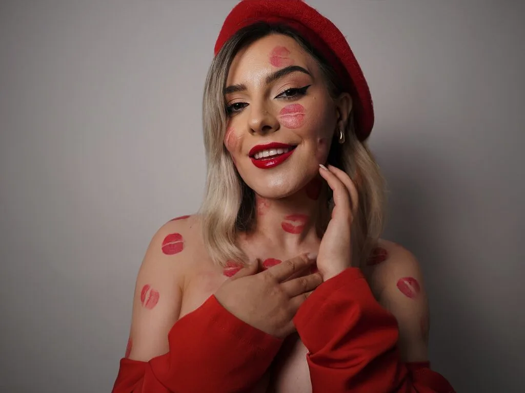 JessieBond smiles with red lipstick on her lips and red kiss marks across her face, neck, and shoulders while wearing a red beret.