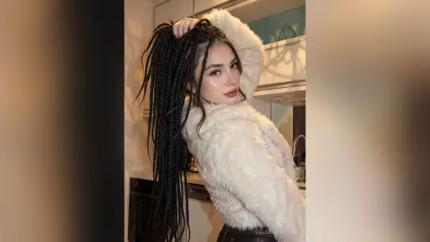 Young woman with long black braids wearing a cozy white faux fur jacket in a modern kitchen setting.
