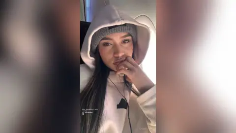 Young woman in a cozy white hoodie and grey beanie looking thoughtful in a bright indoor setting.