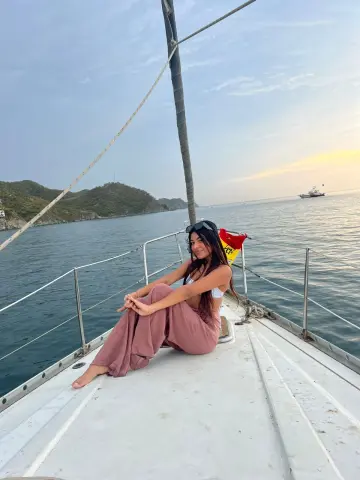 Smiling woman in a white bikini and pink trousers relaxing on a sailboat deck at sunset.