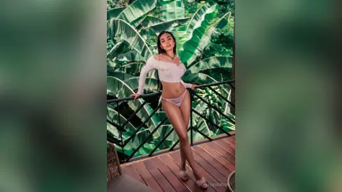 Young woman in white crop top and lace bikini bottoms posing on a tropical wooden balcony surrounded by lush greenery.