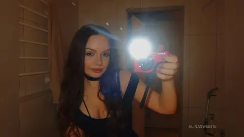 Young woman in a black outfit taking a moody selfie with a bright camera flash in a dimly lit bathroom.