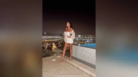 ArianaKroft stands on a rooftop terrace at night wearing a short white dress and heels against a city skyline view.