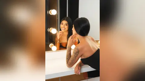 Smiling woman in a black strapless dress looking in a vanity mirror with warm glowing lights.