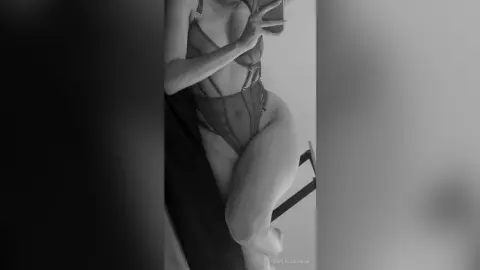 Woman in black lace bodysuit posing for a mirror selfie in a minimalist indoor setting.