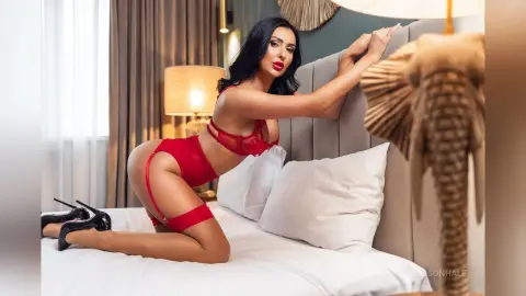 Brunette woman in red lace lingerie and black heels posing on a white bed in a moody bedroom setting.