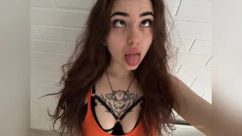Young woman with long wavy brown hair wearing an orange tank top and black harness in a bright indoor setting.