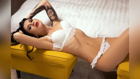 Woman in white lace lingerie relaxing on a yellow velvet ottoman in a bright bedroom setting.