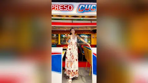Smiling woman in a white floral maxi dress standing inside a colorful vintage food truck.