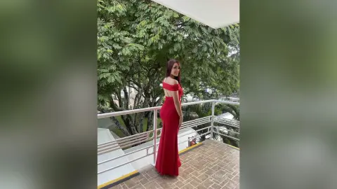 KimRece stands on a balcony wearing a long, off-the-shoulder red dress against a backdrop of green trees.