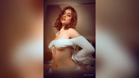 MelisaHank poses with her hair down, wearing a white off-the-shoulder fuzzy top that exposes her breasts and midriff.
