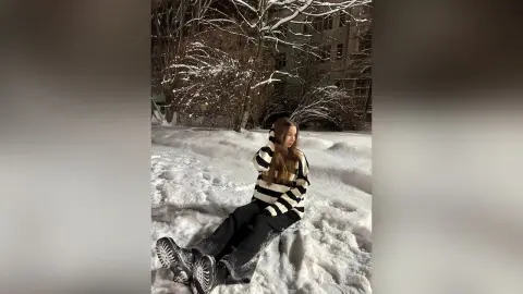 MagalyBoffa sits in the snow wearing a black and white striped sweater, black pants, and black boots with long brown hair.